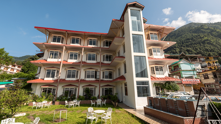 A resort in Bhagsu with a white and red facade, surrounded by outdoor seating in a garden area. The backdrop features green hills and nearby buildings - DLS Meghavan Resort, Dharamshala