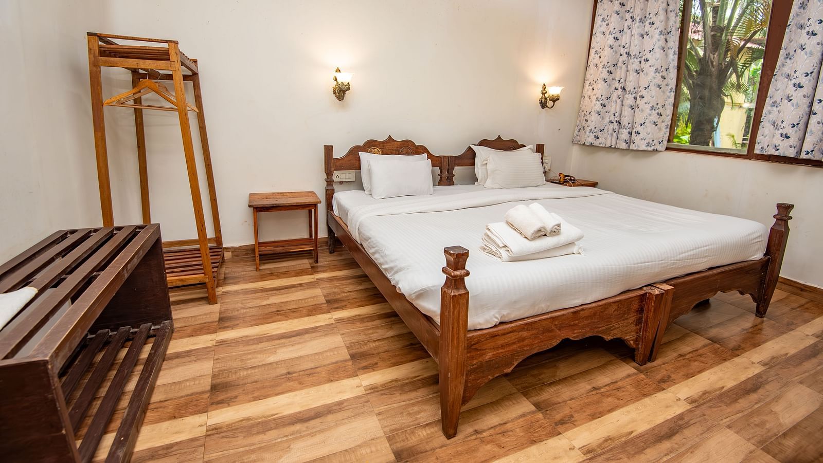 Bright bedroom with a large wooden bed, side lamps, open window, and a luggage rack at De Soul Santé, Morjim.