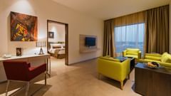 An image of the separate living room of the Suite with sofa set, centre table, work desk, chair, flat screen TV, and an expansive window with curtains - Pride Elite Phaltan