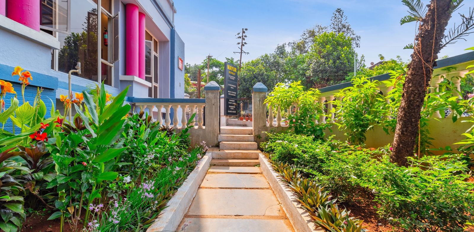 A pathway at Mayur Retreat surrounded by plants and trees, besides the hotel building.