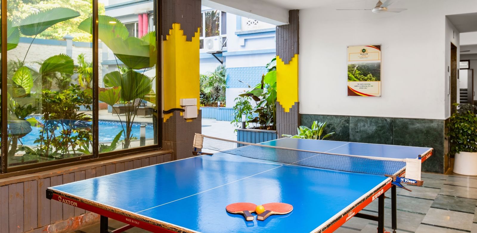 Indoor recreational room at Mayur Retreat featuring a table tennis setup, potted plants, and glass walls offering a mix of fun and relaxation in a lively environment.