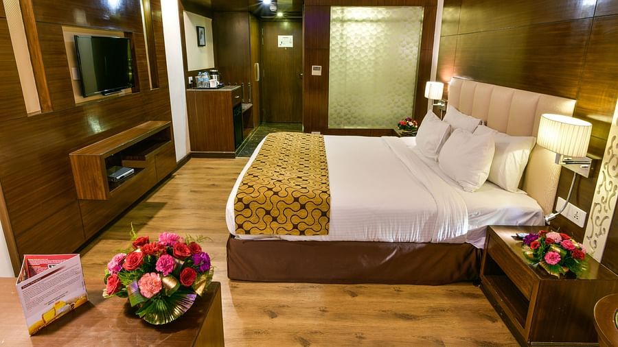 A well-appointed hotel room with wooden accents features a bed with multiple pillows, bedside tables with lamps and flowers, a television, and a glimpse into an adjoining area - Renest Jaipur