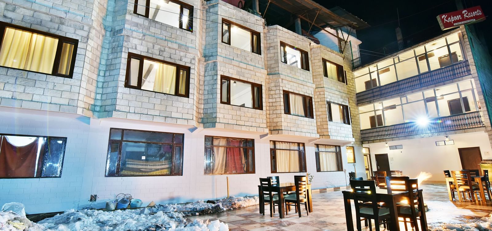 The exterior of DLS Kapoor Resorts Manali at night, showcasing its stone facade, outdoor dining area, and warm lighting.