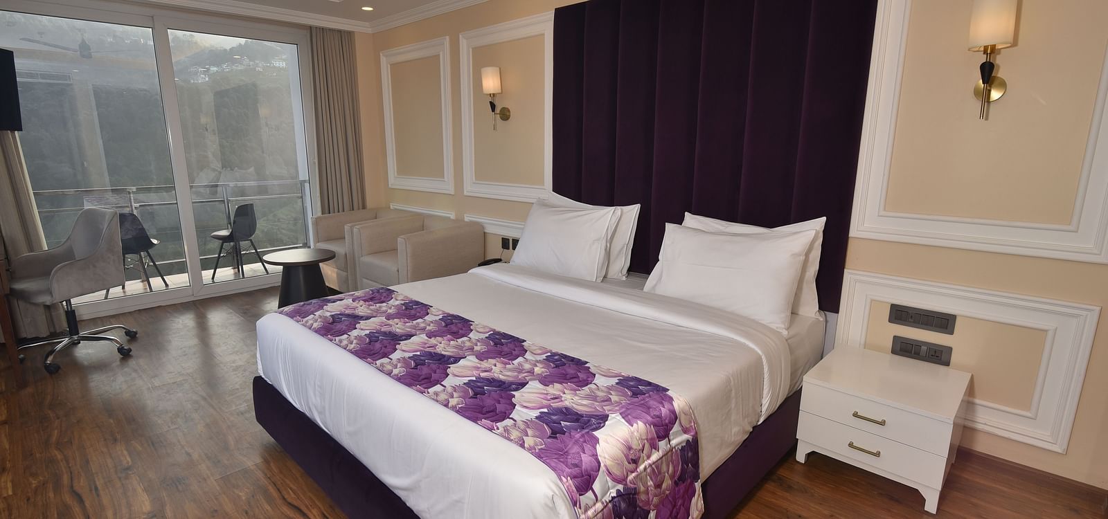 A well decorated modern bedroom. The room features a king-sized bed, a bedside table, a sitting area, and a balcony with outdoor seating. 