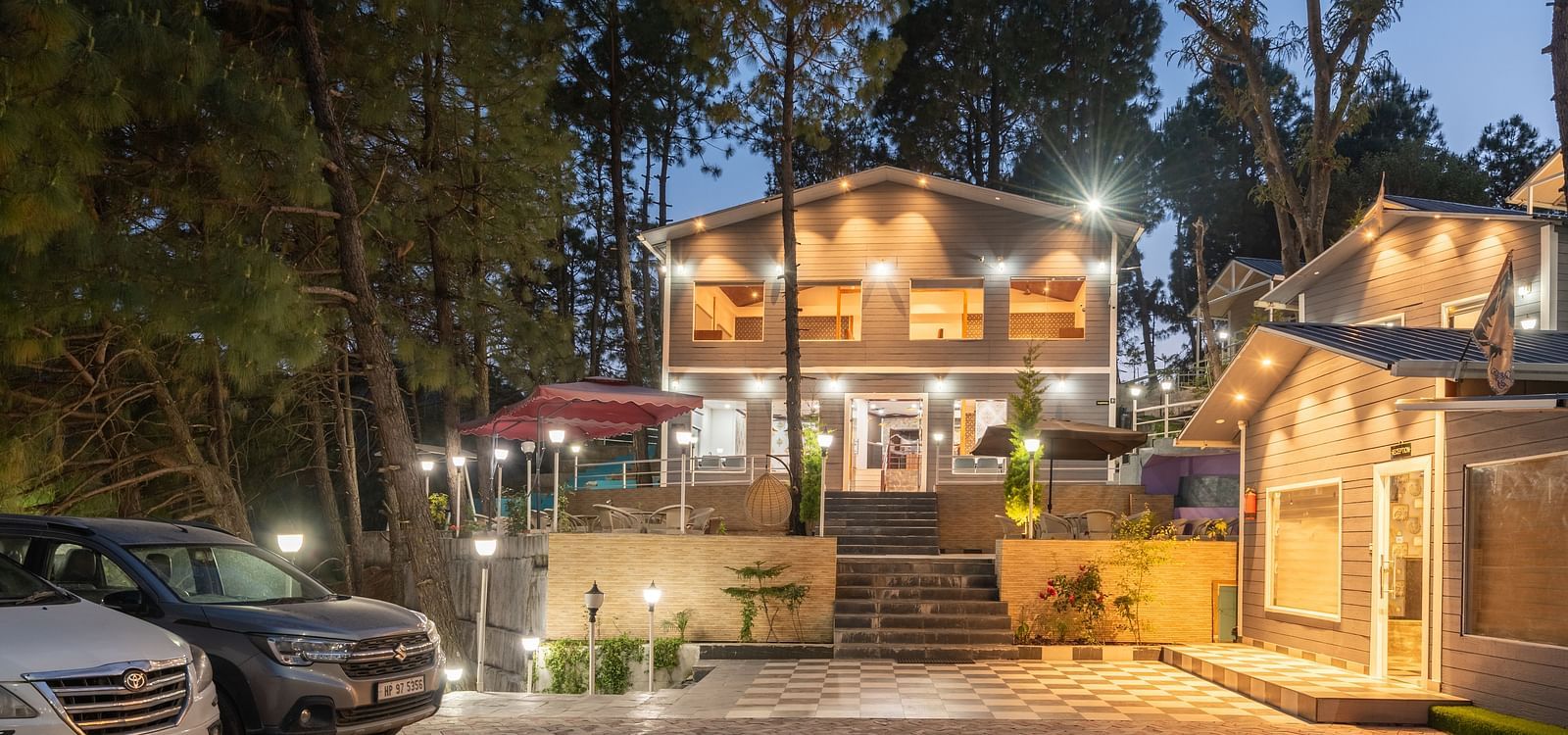 Evening view of a well-lit resort entrance with parked cars, a central staircase leading to a two-story building, and warm ambient lighting surrounded by tall pine trees - DLS Nature Trinket Resort, Dalhousie.