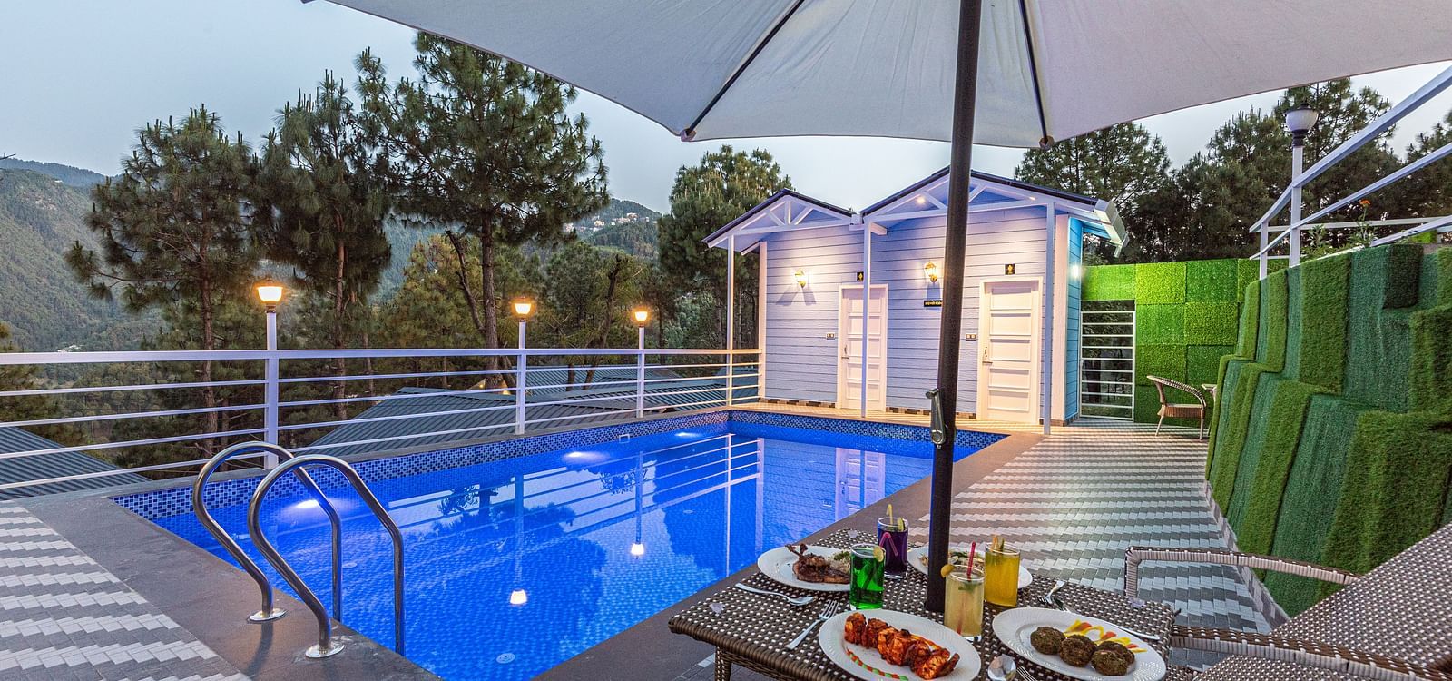 Evening poolside scene with a lit swimming pool, two lounge chairs under a large umbrella, and plates of food and drinks arranged on a table, set against a backdrop of trees and mountains - DLS Nature Trinket Resort, Dalhousie.