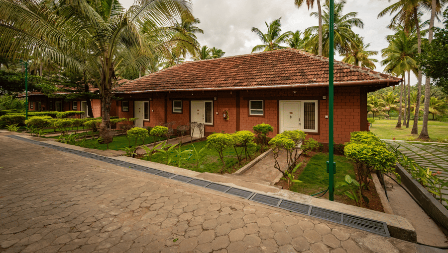 Facade of the River View Suite with gardens, a pathway, and coconut trees under a cloudy sky at Ibex River Resort, Pollachi, one of the best hotels near Aliyar Dam.