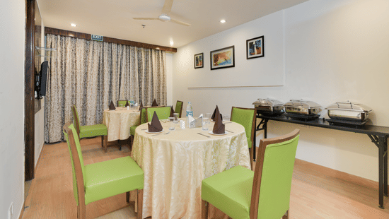 A small, brightly lit dining area at Hotel Bluestone Prime (Nehru Place), New Delhi featuring round tables with light green upholstered chairs and a buffet counter.