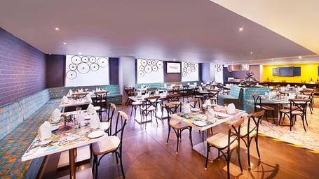 A brightly lit restaurant with chairs, a long bar counter, and a colorful wall mural | Regency Tirunelveli By GRT Hotels