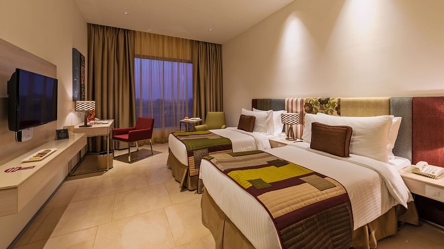 An image of a deluxe twin room with plush beds, bed side table, flat screen TV, lamps, work desk, chair, and decorative items - Pride Elite Phaltan