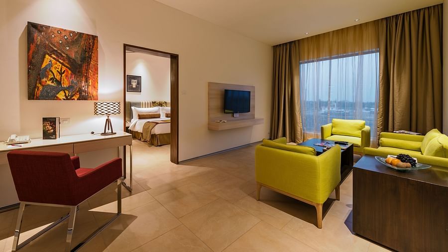 An image of the separate living room of the Suite with sofa set, centre table, work desk, chair, flat screen TV, and an expansive window with curtains - Pride Elite Phaltan