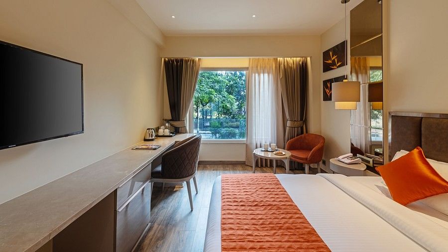 A side view of a double bed room with pillows, sheets, TV, work desk, chairs and an open window with curtains at Hotel Saket 27, New Delhi