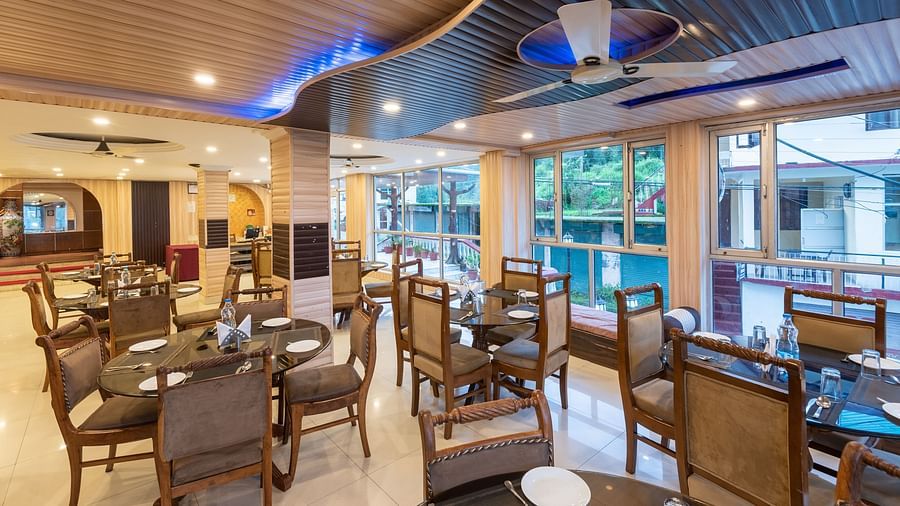 A spacious and restaurant with wooden tables and chairs, a high ceiling with decorative lighting, and large windows overlooking a garden | DLS Anupam Resort, Dharamshala