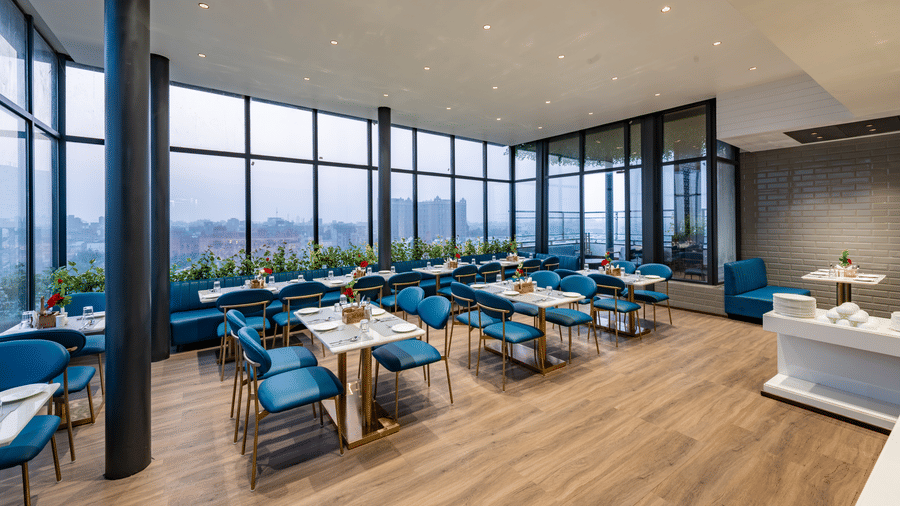 A restaurant with arranged tables, chairs, large windows, ceiling lights, and food counter at DLS Hotels Amritsar.
