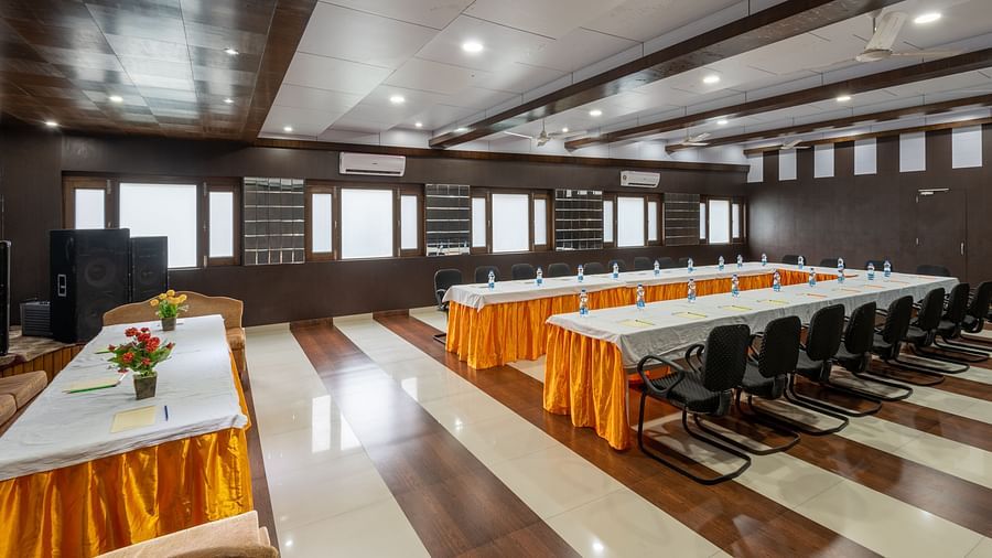 A conference room with a long table, chairs, and decorative flowers. The room has wooden panelling, bright lighting, and windows along the walls - DLS Meghavan Resort, Dharamshala