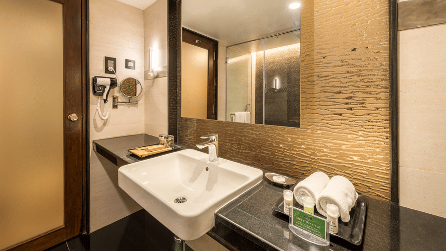 Club Room bathroom vanity at The Citrine, Bangalore, with a white basin, large mirror, gold textured wall panel, and folded towels on a black counter.