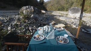 A table set with 2 plates and drinks under a canopy next to a rocky riverbed with hills.