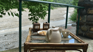 A teapot, 2 cups and snacks on a balcony table overlooking a wide river and green hills.