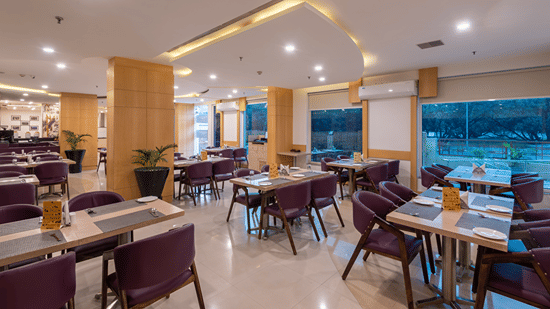 Restaurant dining area with tables and chairs beside large windows at Mango Grove Hotel, Chandigarh.
