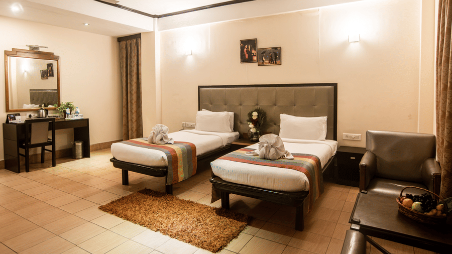 A well-lit hotel room at Polo Tura Hotel with twin beds, neutral decor, seating area, a work desk with a mirror, and neatly arranged linens.
