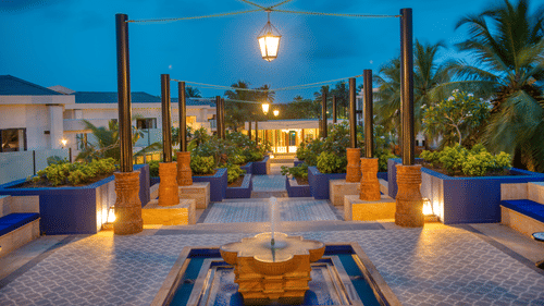 A wide, symmetrical stone pathway leads through a coastal resort towards a central pavilion. Multi-level planters filled with lush tropical vegetation line the walkway. Palm trees and contemporary buildings flank the path under a sunset sky with clouds.