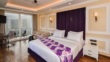 A vibrant bedroom with a large bed featuring purple accents and a bright atmosphere. The room includes large windows that provide scenic views of the hills outside.