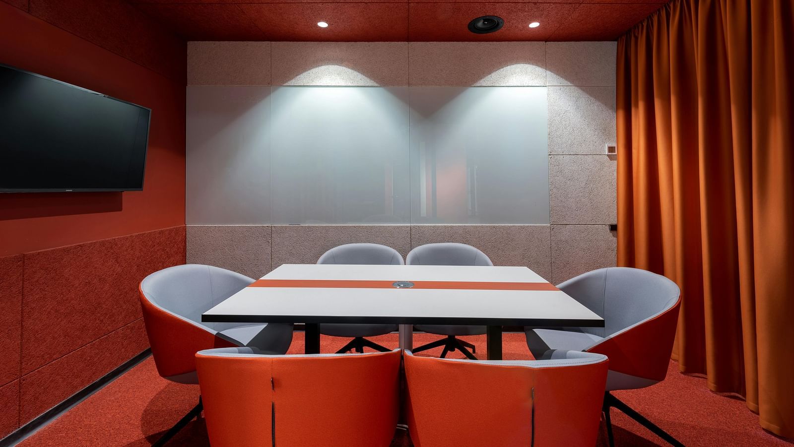 A modern conference room with red walls, a white table surrounded by grey and orange chairs, and a large screen on the left wall.