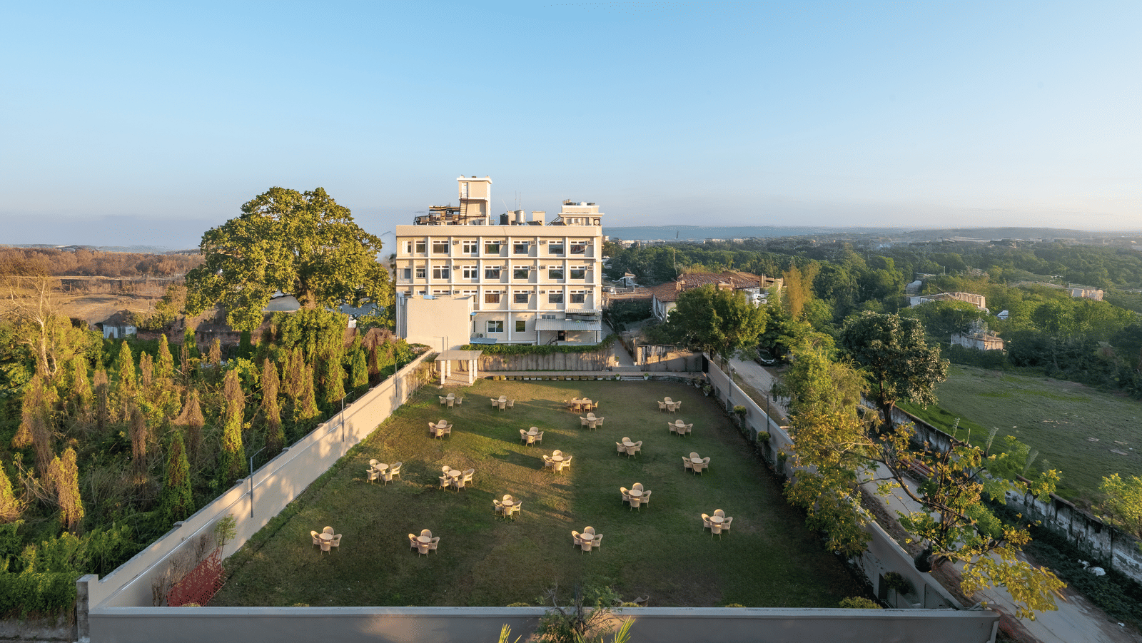 An aerial view of the lawn area at DLS Park Grace, Haridwar, one of the best Wedding Resorts in Haridwar, featuring several sets of tables and chairs surrounded by luscious green trees and site of the clear blue sky.