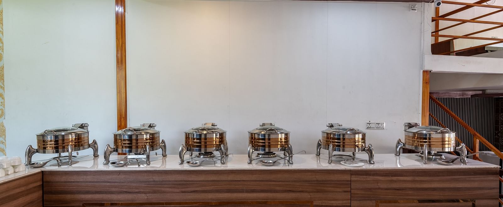  Buffet setup with stainless steel chafing dishes arranged on a wooden counter against a plain wall in a modern restaurant - DLS Nature Trinket Resort, Dalhousie.