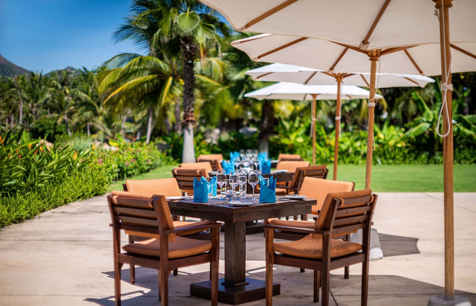 A coastal outdoor dining space with wooden furniture, white sunshades, amidst palm trees and tropical foliage - Alibu Resorts Nha Trang