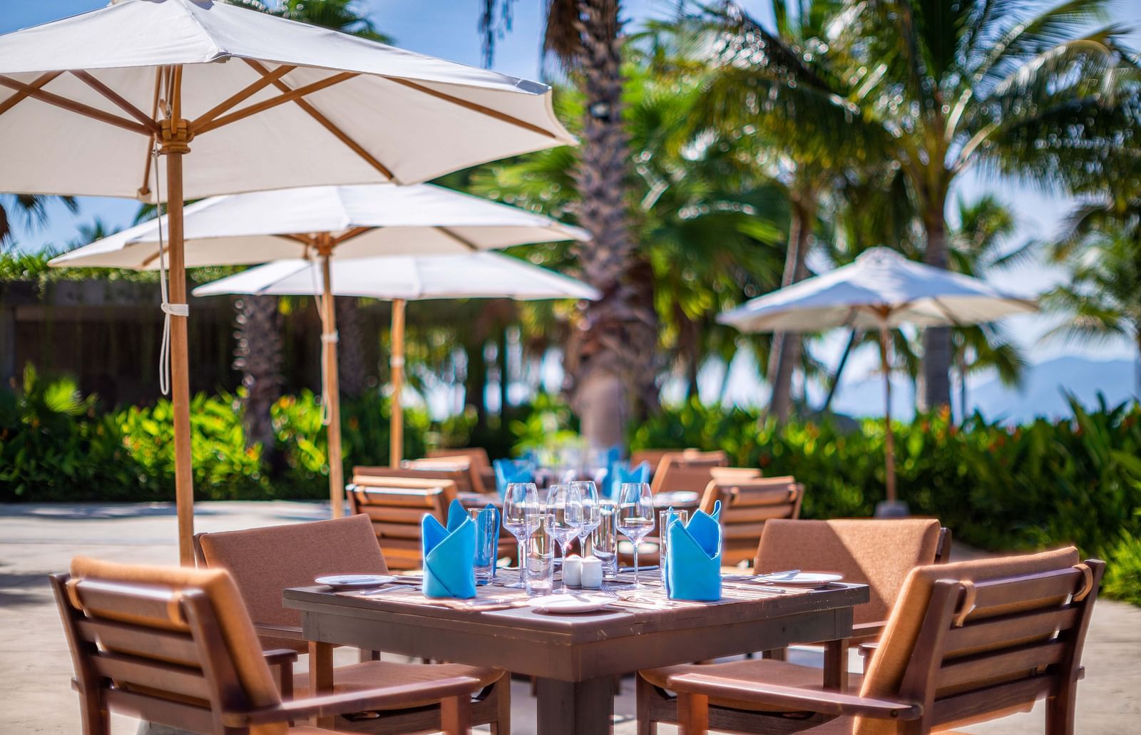 A picturesque dining setting under white umbrellas with set tables, palm trees and a lush background - Alibu Resorts Nha Trang