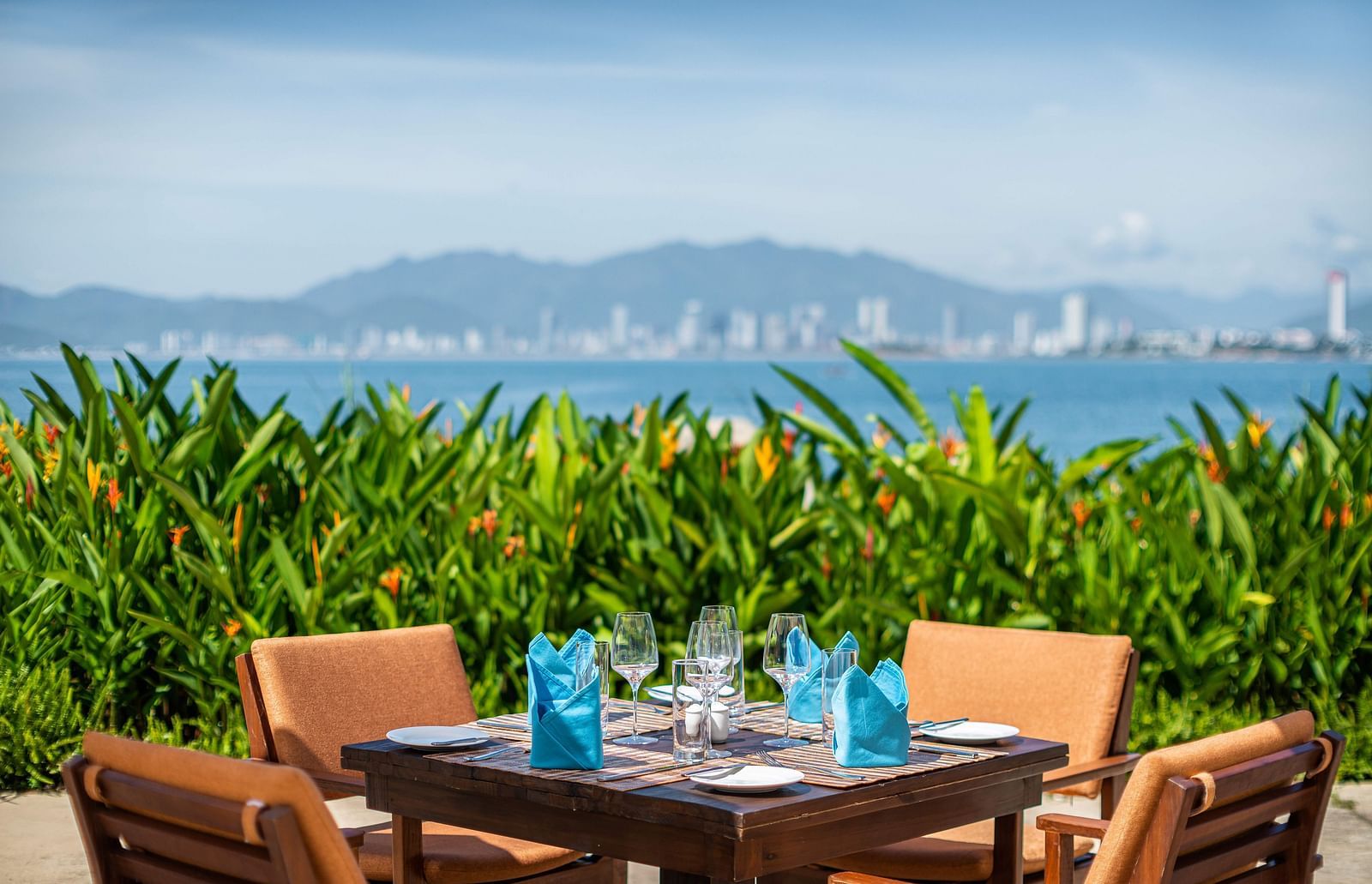 A seaside dining area with set tables under white umbrellas, vibrant plants and a panoramic view of the coastline and city - Alibu Resorts Nha Trang