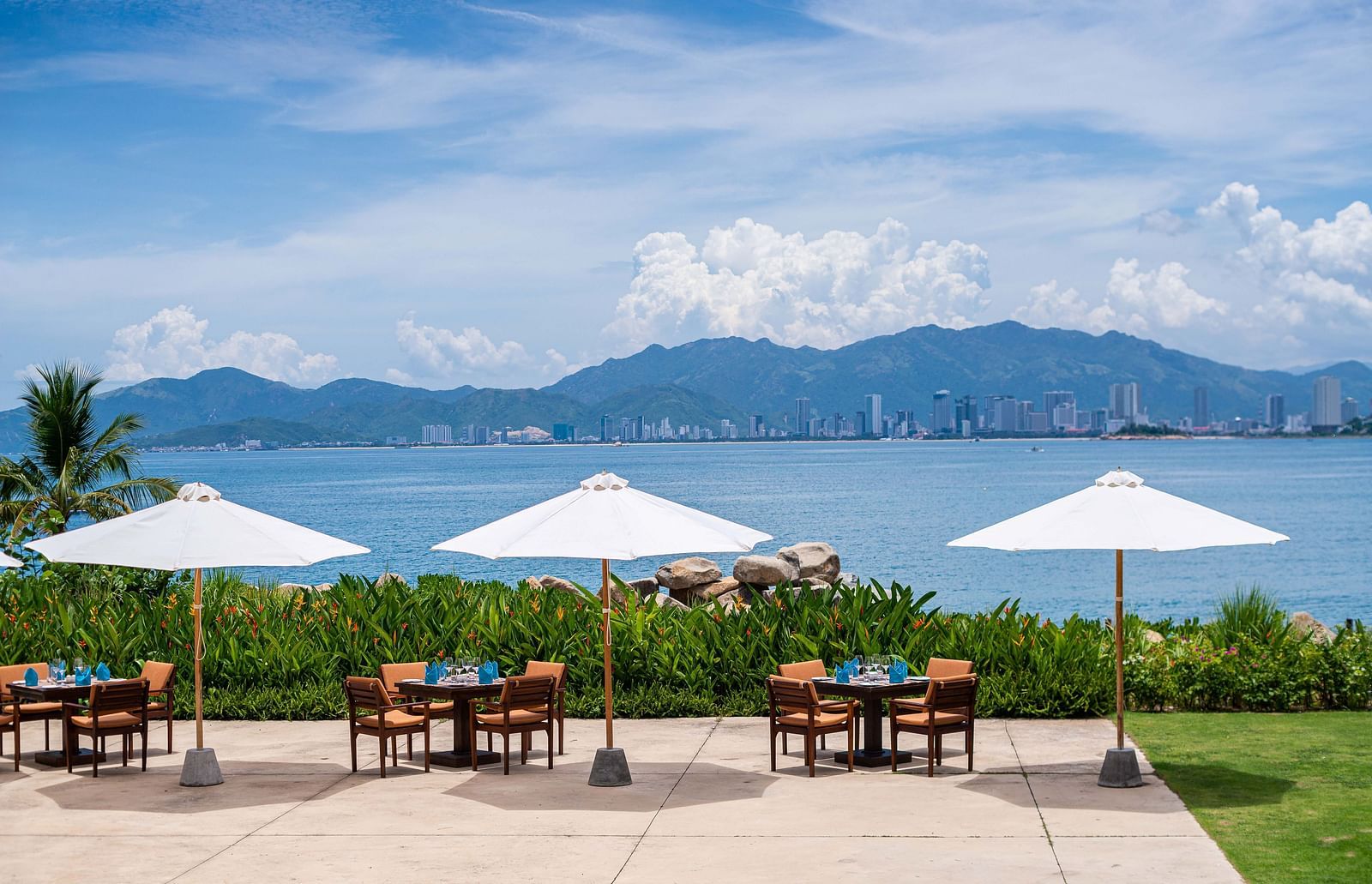 An alfresco dining setup with white umbrellas overlooking a serene bay and a distant cityscape - Alibu Resorts Nha Trang