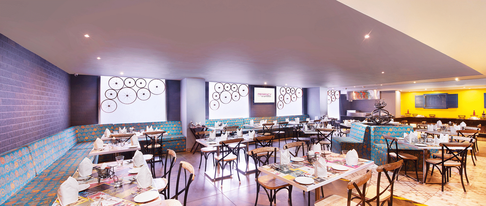 A brightly lit restaurant with chairs, a long bar counter, and a colorful wall mural | Regency Tirunelveli By GRT Hotels