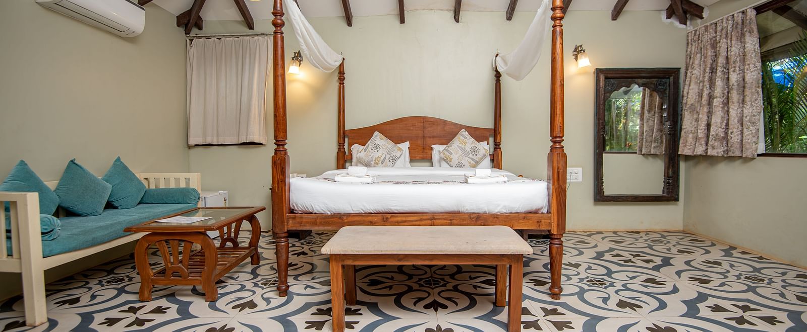 Spacious room with a large four-poster bed, cushioned sofa and wooden table, and a side window with curtains pulled aside at De Soul Santé, Morjim.