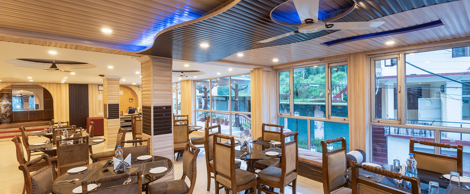 A spacious and restaurant with wooden tables and chairs, a high ceiling with decorative lighting, and large windows overlooking a garden | DLS Anupam Resort, Dharamshala