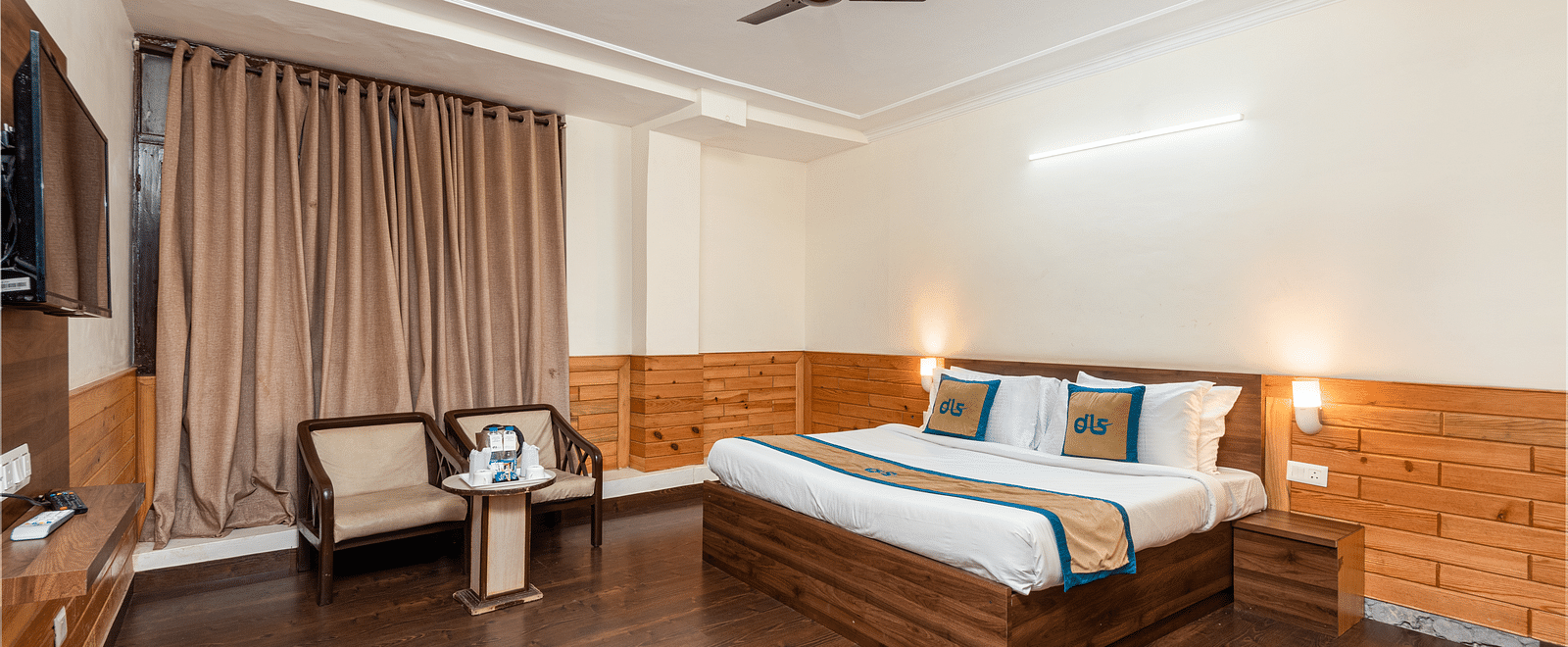 A well lit hotel room with a wooden floor, a large bed, two armchairs, a small table, and a ceiling fan | DLS Anupam Resort, Dharamshala