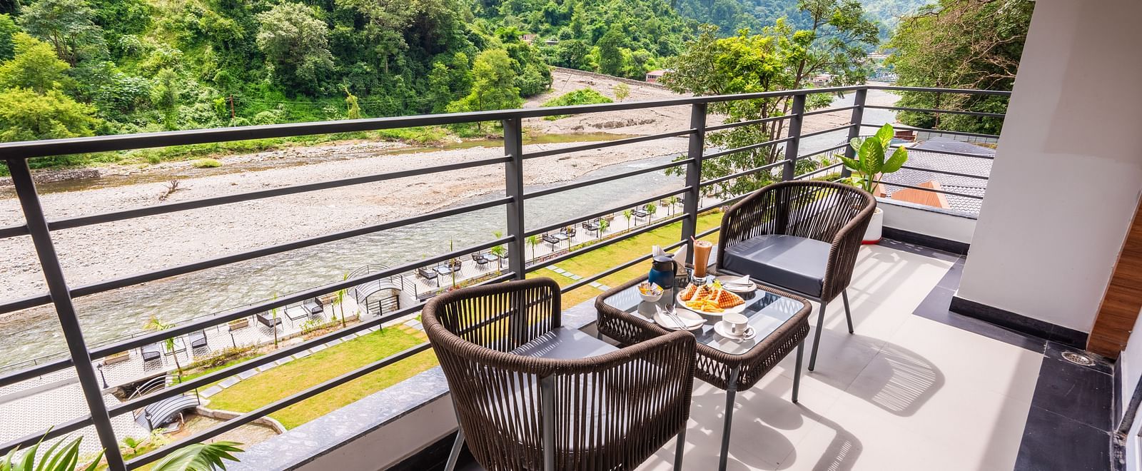 Overview of balcony of Premium Room at DLS Divine River Resort and  Spa featuring seating area with food kept on table and backdrop of river and forest views.