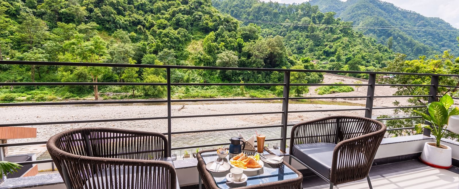 Balcony of Premium Room at DLS Divine River Resort and  Spa featuring seating area with food kept on table and backdrop of river and forest views.