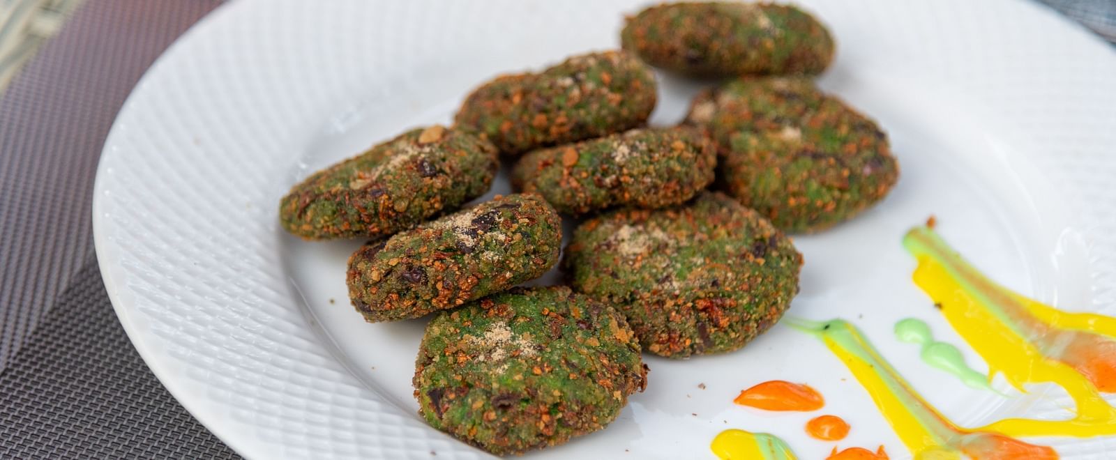 Close-up of a white plate with a neatly arranged serving of green vegetable cutlets, garnished with colourful sauce drizzles on the side - DLS Nature Trinket Resort, Dalhousie.