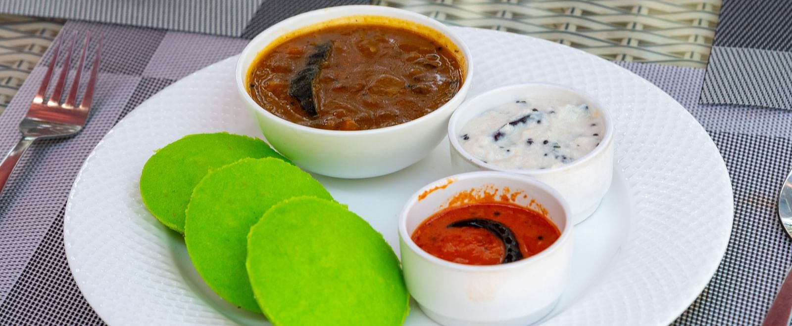 A South Indian breakfast plate with three green idlis, served with bowls of sambar, coconut chutney, and tomato chutney on a white plate at DLS Nature Trinket Resort, Dalhousie.