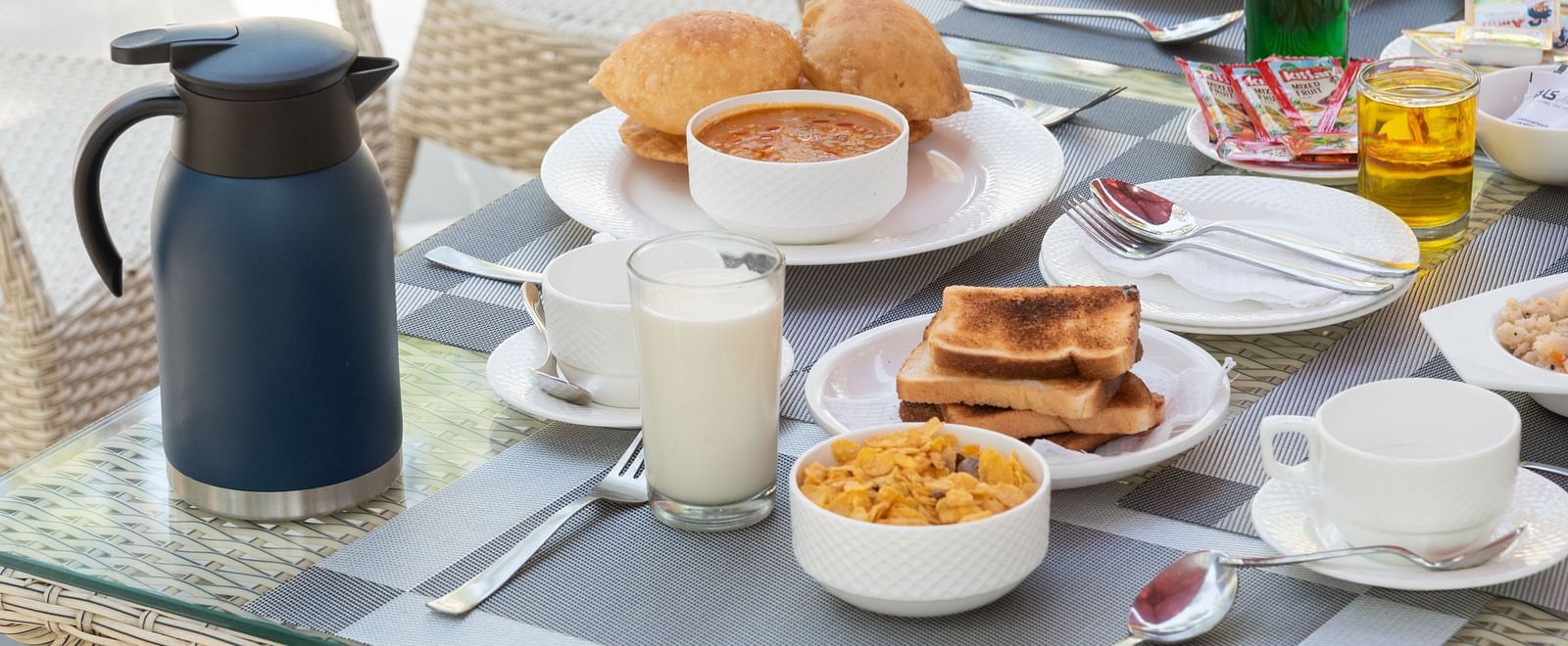 Close-up of a breakfast table set with cereal, toast, buns, tea or coffee, juice, and condiments on a glass-top table with wicker chairs - DLS Nature Trinket Resort, Dalhousie.