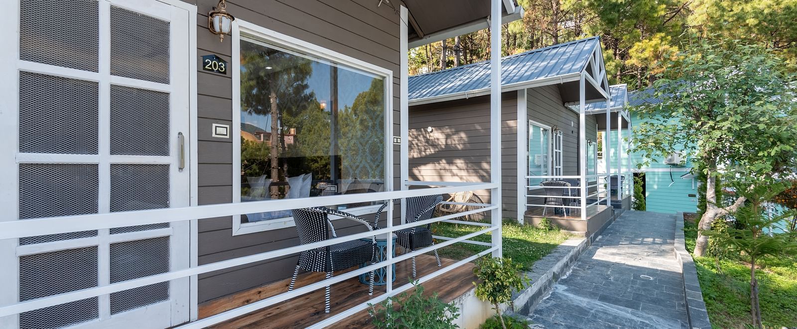 Row of modern wooden cottages with front patios, white railings, and a stone pathway surrounded by greenery and tall pine trees - DLS Nature Trinket Resort, Dalhousie.