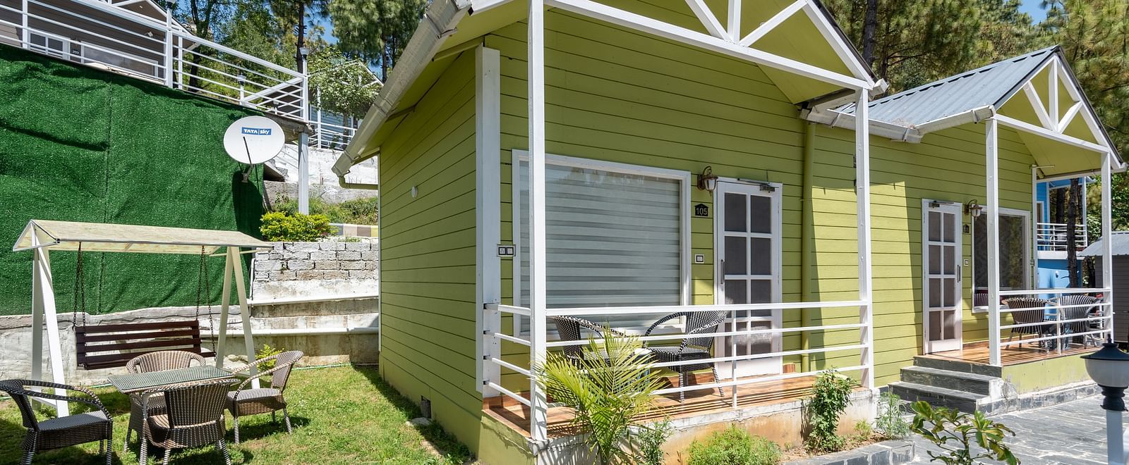 Bright green wooden cottages with white trim, set along a stone pathway, featuring a grassy outdoor seating area with a swing and surrounded by tall trees - DLS Nature Trinket Resort, Dalhousie.