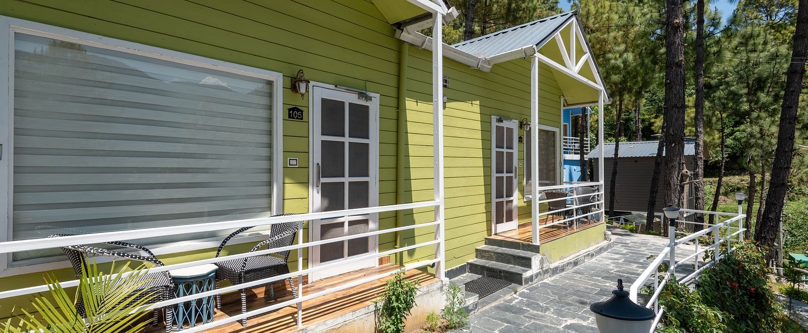  Row of vibrant green wooden cottages with white railings along a curved stone pathway, nestled among tall pine trees in a serene forested setting - DLS Nature Trinket Resort, Dalhousie.