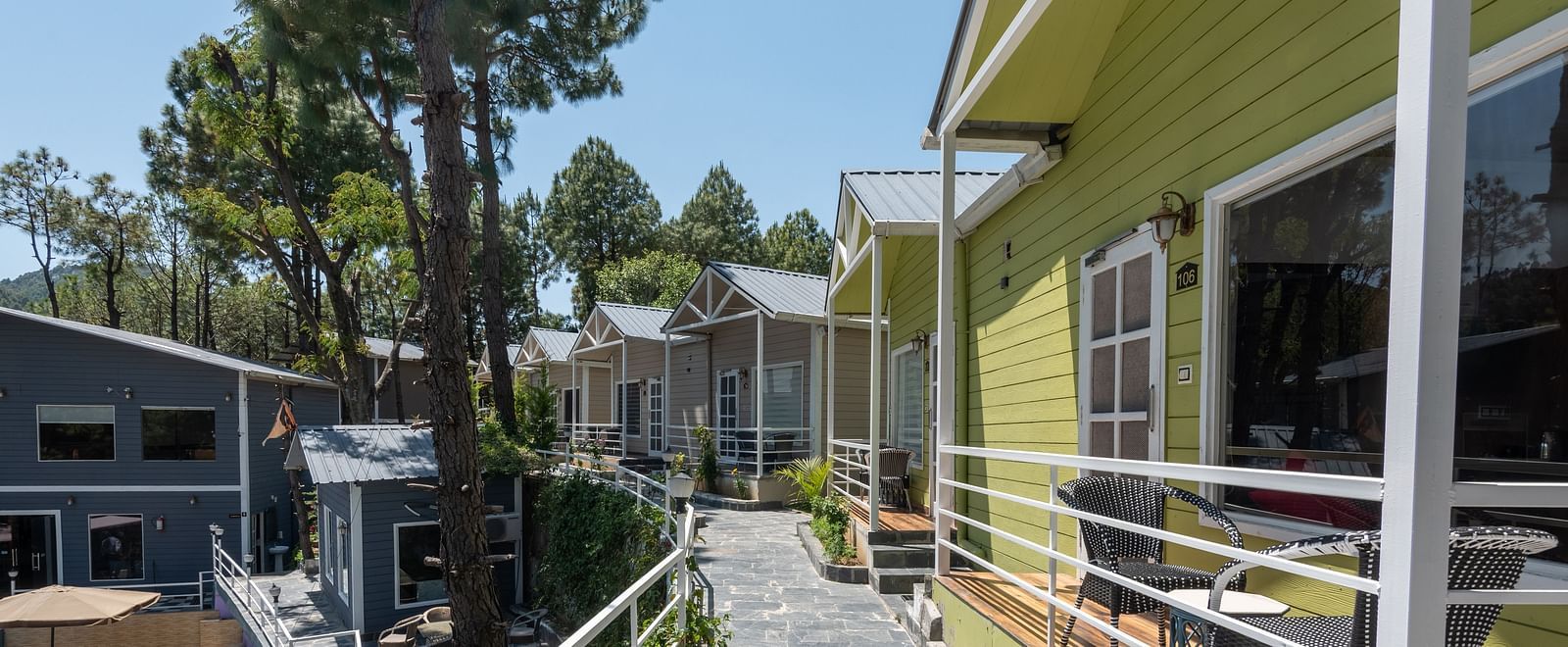 Stone-paved pathway running alongside green wooden cottages with white railings, outdoor seating areas, and surrounded by tall pine trees in a hilly setting - DLS Nature Trinket Resort, Dalhousie.