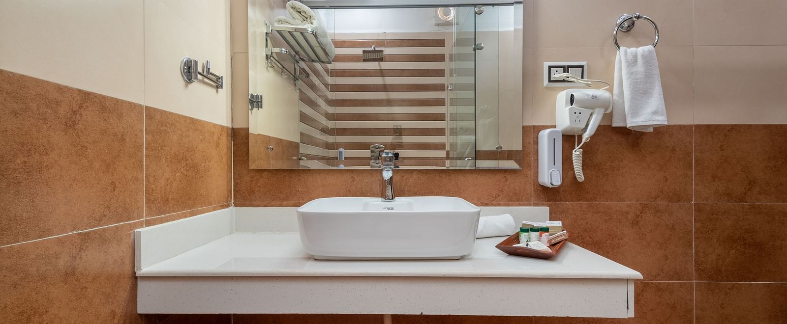 A modern bathroom vanity area featuring a square white basin atop a sleek countertop, with a wall-mounted mirror, a hairdryer, towel ring, and a rack with neatly arranged shelves reflected in the background - DLS Nature Trinket Resort, Dalhousie.