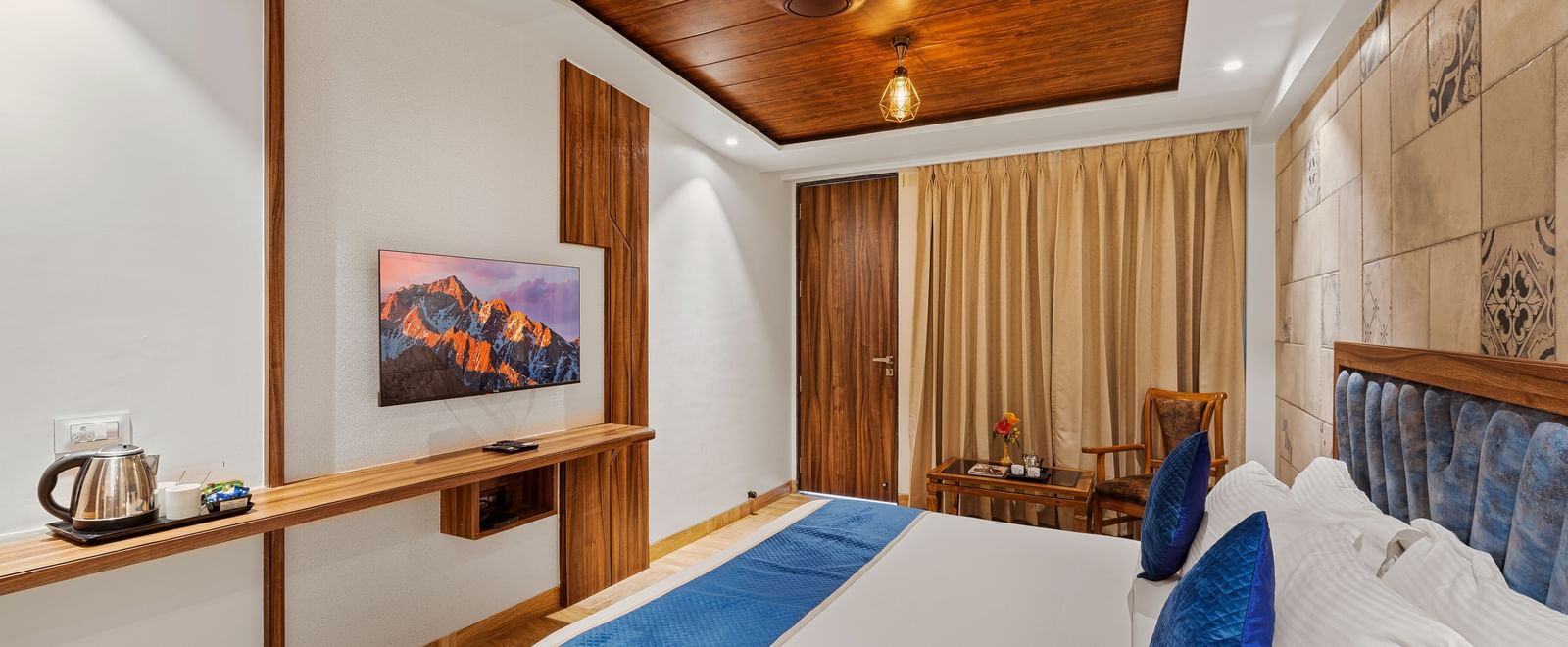 Spacious bedroom with a blue-accented bed, wooden decor, and curtains drawn over large windows at Mayur Retreat.