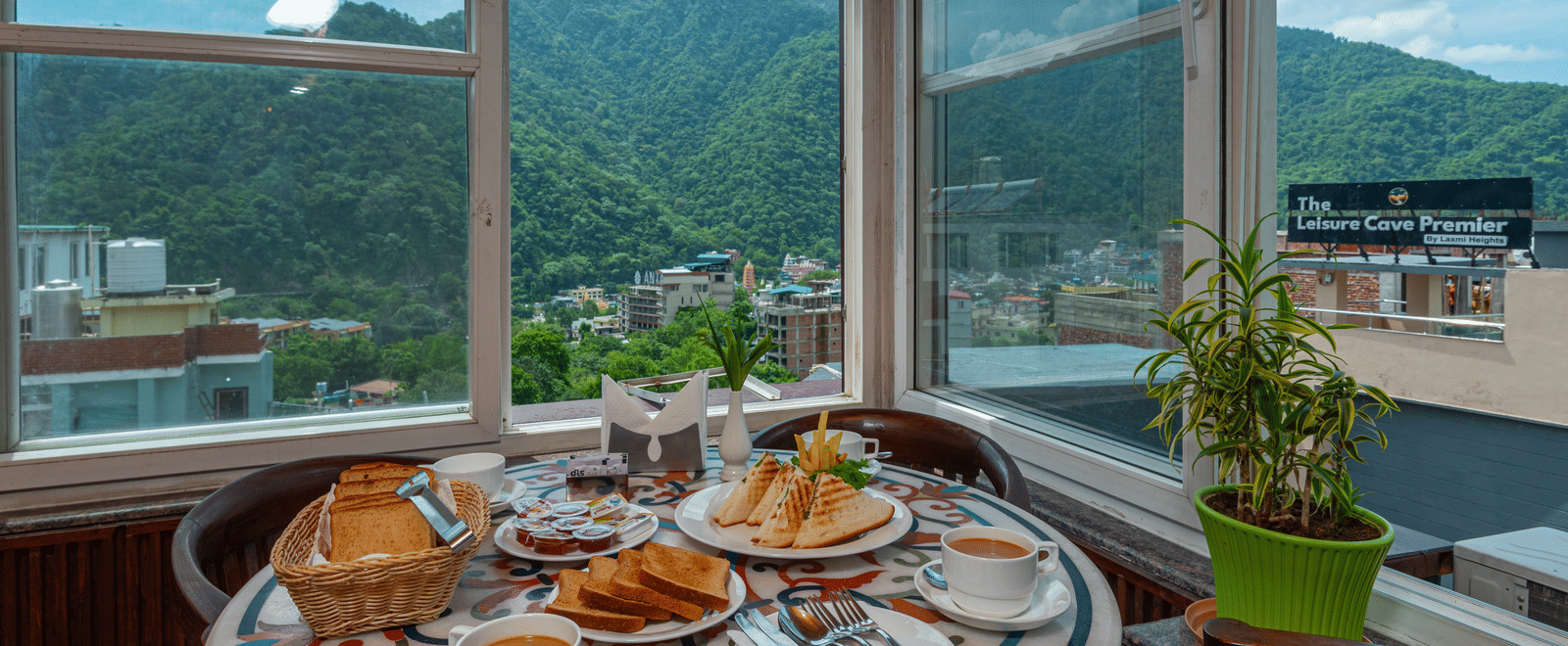 A cafe table is set for breakfast, with plates of toast, grilled sandwiches, jam, and coffee. The setting is bright with natural light from large windows, offering a view of a green, forested mountain and a townscape below. This is a shot from the cafe at 