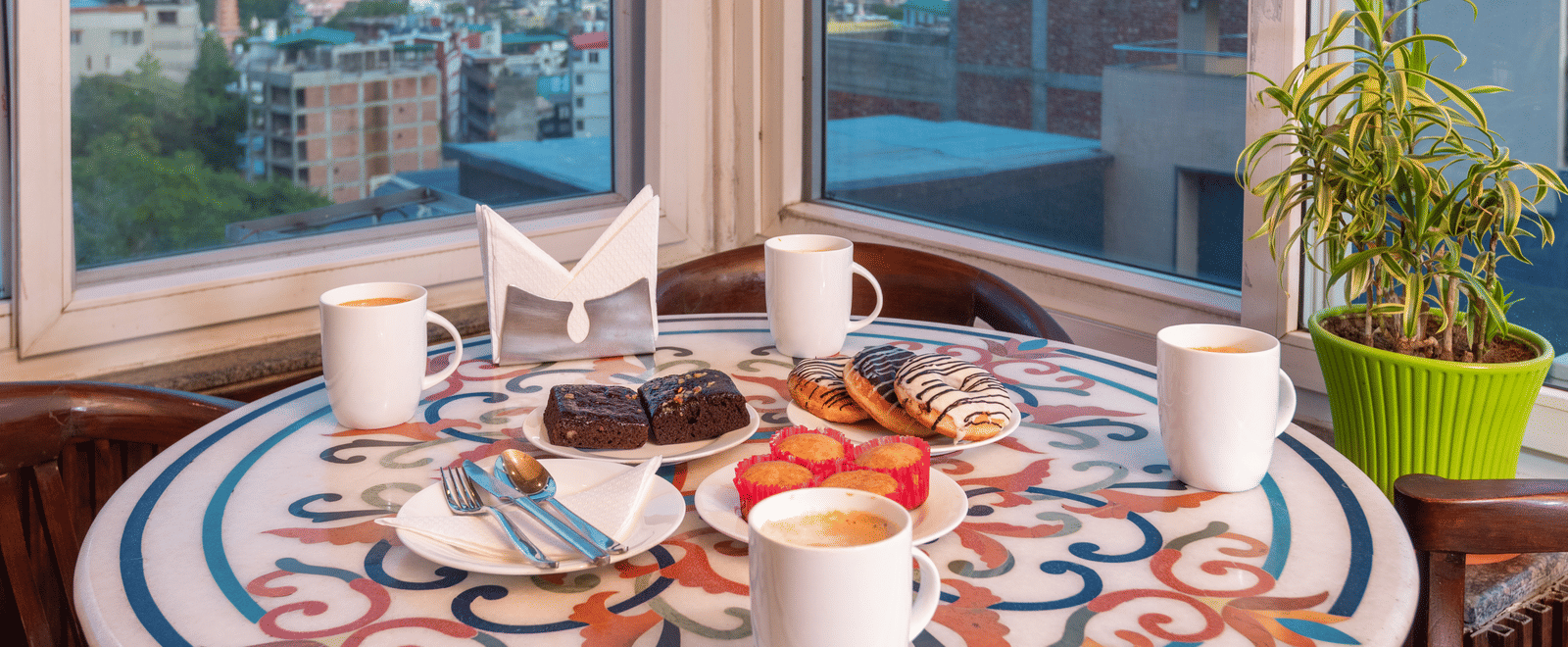 A cafe table is set with a selection of pastries, donuts, and four cups of coffee. The table is decorated with a colorful, round mosaic pattern. In the background, a large window reveals a view of the city and surrounding green hills. This is a shot of the